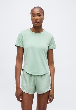 Mujer con camiseta de manga corta verde claro y pantalones cortos deportivos a juego, de pie frente a un fondo blanco liso.