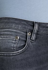 Jeans gris oscuro con un acabado liso, que presentan bolsillos frontales, remaches metálicos y un lazo para cinturón. Costuras cosidas con un ligero detalle de desvanecimiento.