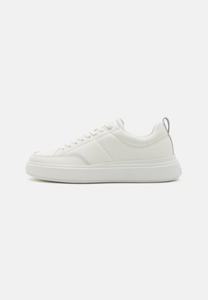 Hvid sneaker med en glat læderoverdel, rund tå, snørebånd foran og en kontrastfarvet grå trækflap bagpå. Tykk hvid gummisål.