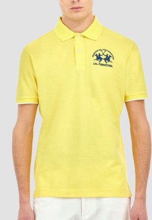 Mężczyzna ubrany w żółtą koszulkę polo z krótkim rękawem i niebieskim haftowanym logo na lewej piersi oraz białe spodenki.
