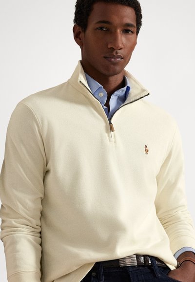 Muž ve smetanovém svetru na čtvrtý zip přes světle modrou košili s límečkem, tmavé džíny a hnědý opasek, stojící před bílým pozadím.