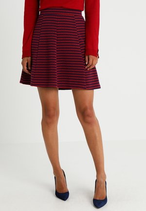 Femme portant une jupe évasée rayée rouge et bleu marine, associée à des talons hauts bleu marine et un haut rouge à manches longues, debout devant un fond uni.