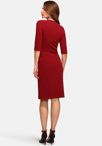 Rotes, figurbetontes Kleid mit Dreiviertelärmel, knielang, glattes Material, dezenter Akzent in der Taille, getragen mit schwarzen hohen Schuhen.