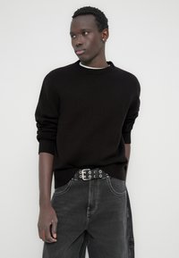 Μαύρο πλεκτό πουλόβερ με στρογγυλή λαιμόκοψη, σετ με σκούρο γκρι τζιν και μαύρη ζώνη με ασημένιες λεπτομέρειες και τρουκ.