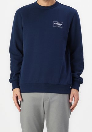 Mand iført en mørkeblå Tommy Hilfiger sweatshirt med logo på brystet og lysegrå bukser, stående mod en hvid baggrund.