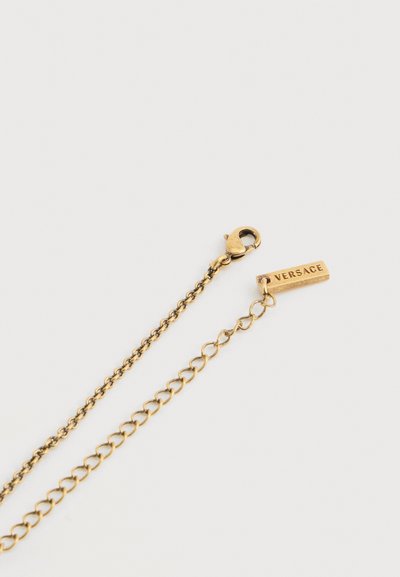 Collier chaîne en or avec fermoir mousqueton et étiquette rectangulaire gravée de "VERSACE" sur fond blanc.