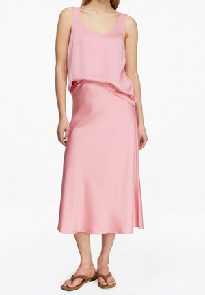 Woman wearing a sleeveless pink satin top and matching midi skirt with brown flat sandals, standing against a plain white background.