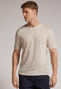 Beige tričko s krátkým rukávem vyrobené z jemného, texturovaného materiálu s kulatým výstřihem, v kombinaci s tmavě modrými kalhotami. Viditelný jemný melírovaný vzor.