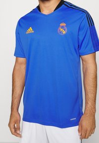 Královsky modrý dres Adidas s designem do véčka, černými akcenty na ramenou, se znakem Real Madrid v oranžové barvě na hrudi.
