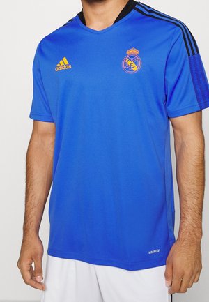 Royalsininen Adidas-paita, jossa on V-pääntie, mustat yksityiskohdat hartioissa, ja jonka rinnassa on oranssi Real Madrid -logo.