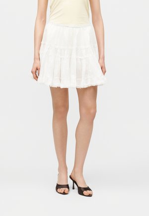 Mini-jupe blanche à volants portée par une personne avec un haut beige et des mules à talons ouvertes noires, sur un fond clair uni.