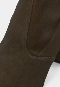 Gros plan sur une bottine en daim marron avec panneau latéral élastique et talon carré sur fond blanc.