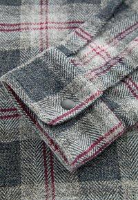 Cămașă gri cu carouri și accente roșii, cu un model în țesătură herringbone. Fabrică din bumbac, închidere cu nasturi la mansetă, detalii cu țesătură texturată.