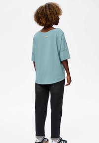 Lichtblauw oversized t-shirt met wijde halslijn en korte mouwen, gecombineerd met donkere spijkerbroek. Heeft een klein logo op de achterkant.