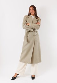 Cappotto beige doppiopetto con ampi rever e cintura. Il tessuto ha una texture liscia. Abbinato a pantaloni crema e tacchi neri.