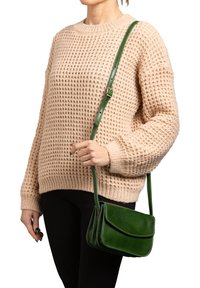 Donna che indossa un maglione beige a maglia e pantaloni neri, porta una borsa a tracolla in pelle verde con tracolla regolabile.
