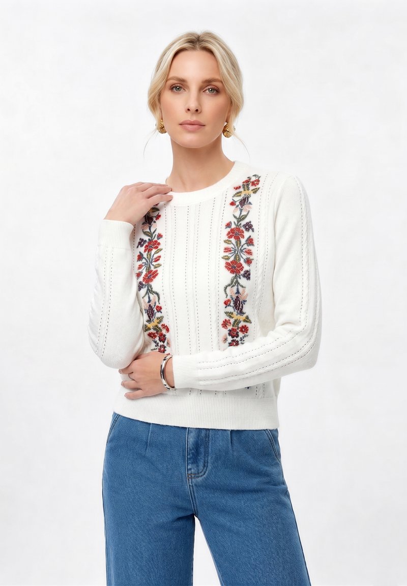 Femme aux cheveux blonds portant des boucles d'oreilles en or, un pull blanc avec une broderie florale verticale et un jean bleu, posant devant un fond uni.