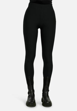 Persona che indossa leggings neri aderenti abbinati a stivali neri in pelle robusta, in piedi davanti a uno sfondo chiaro e uniforme.
