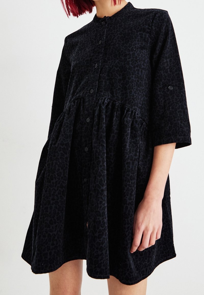 Femme portant une robe noire ample à boutons avec un imprimé léopard subtil et des manches trois-quarts, debout devant un fond blanc uni.