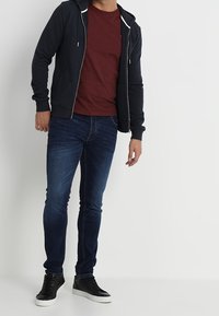 Sweat à capuche zippé coupe athlétique en bleu marine foncé, porté avec un t-shirt bordeaux, un jean slim bleu et des baskets noires.