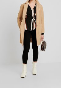 Cappotto in misto lana color cammello con colletto a rotondo, blouse nera a righe, jeans skinny neri e stivaletti bianchi con tacco a blocco. Borsa marrone.