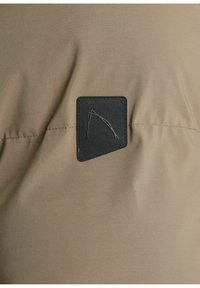 Rjava tkanina z gladko teksturo, ki ima črn pravokoten odstranek z minimalističnim logotipom v zgornjem levem kotu.