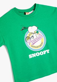 Camiseta verde de manga corta hecha de algodón, con un gráfico de Snoopy sosteniendo una raqueta de tenis y el texto "Departamento de Peanuts" en colores.