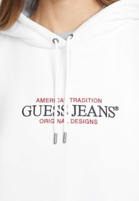 Sudadera con capucha blanca con cierre de cordón y puntas metálicas plateadas. Presenta el bordado "GUESS JEANS®" en negro y rojo en la parte delantera.