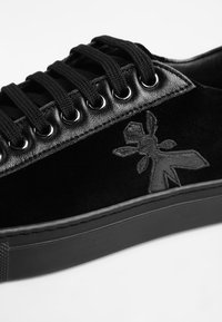 Baskets noires avec lacets noirs, œillets métalliques, garniture en cuir et logo brodé en forme de fourmi sur le côté au-dessus de la semelle.
