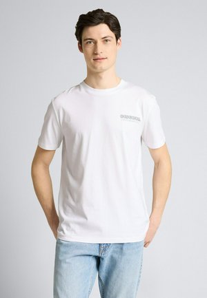 Junger Mann mit heller Haut und dunklen Haaren steht und trägt ein weißes T-Shirt mit dem Schriftzug "ORIGINAL" und hellblaue Jeans vor einem einfarbigen Hintergrund.