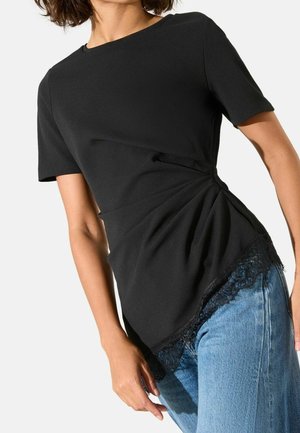 Femme portant un haut noir à manches courtes avec un détail froncé sur le côté et une bordure en dentelle, associée à un jean bleu sur un fond uni.
