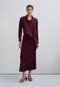 Blusa burdeos con escote drapeado combinada con una falda cruzada a juego. Textura transparente, larga hasta el tobillo, complementada con tacones negros de tiras.