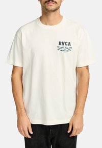 Hvid kortærmet t-shirt lavet af bomuld, med et grafisk print i teal, der viser teksten "RVCA" og "NATURE VERSUS INDUSTRY" på fronten.