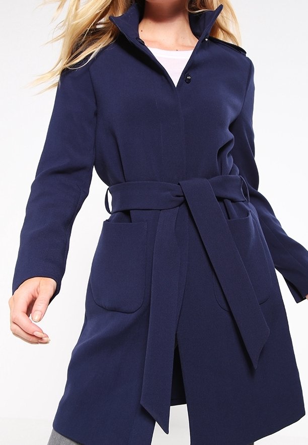 Femme portant un manteau bleu marine ceinturé avec de grandes poches avant et un col montant sur un haut blanc.