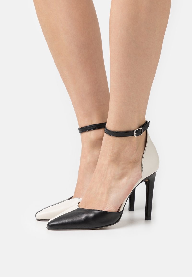 Des chaussures à talons hauts en noir et blanc avec un bout pointu, dotées d'une bride de cheville noire et d'une texture en cuir lisse. Design avec un blocage de couleur distinct.