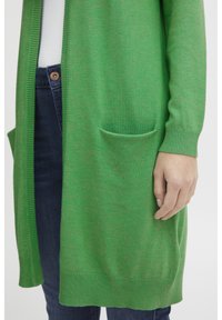 Cardigan en tricot vert avec des poignets et un ourlet côtelés, dotée de poches latérales et d'une coupe décontractée, associée à un jean foncé.