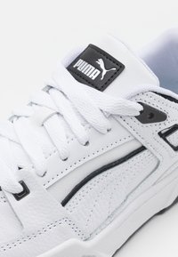 Baskets blanches Puma avec logo noir sur la languette, lacets blancs épais et panneaux en cuir superposés avec détails de garniture noire.