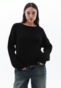 Maglione nero a maglia con scollo ampio, texture a coste e fit morbido. Indossato con jeans blu scoloriti, che mostrano uno stile rilassato.