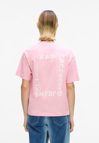 Roosa puuvillane t-särk, millel on seljal valged tekstigraafikad, lühikeste varrukatega ja mugava istuvusega, kombineeritud siniste teksapükstega.