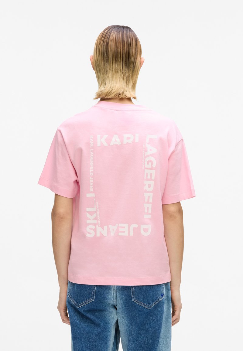 Roosa puuvillane t-särk, millel on seljal valged tekstigraafikad, lühikeste varrukatega ja mugava istuvusega, kombineeritud siniste teksapükstega.