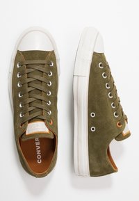Sneakers basse in pelle scamosciata verde oliva con punte e suole in gomma bianca, esposte da vedute dall'alto e di lato su uno sfondo bianco.