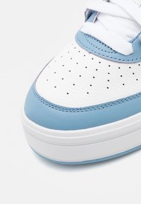 Witte leren sneaker met blauwe accenten, geperforeerde neus, platte witte veters en een dikke witte zool. Glad oppervlak met contrasterende stiksels.