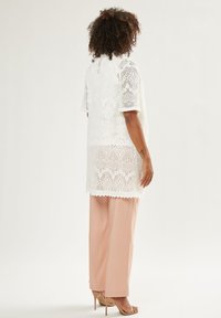 Tunika bianca in pizzo con maniche corte, caratterizzata da motivi intricati e orlo scallop. Abbinata a pantaloni a palazzo rosa chiaro e tacchi beige.