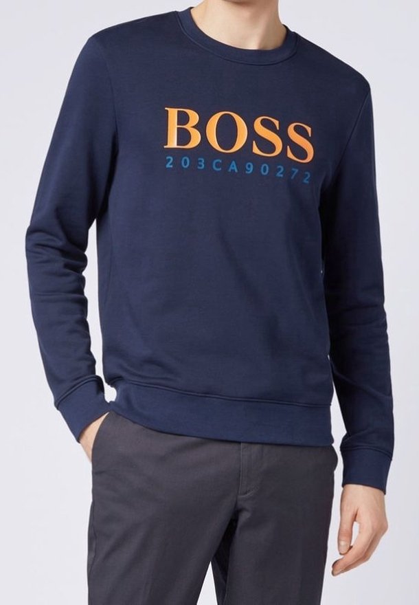 Sweat-shirt bleu marine avec texte orange "BOSS" et chiffres bleus, porté par une personne avec la main dans la poche et un pantalon gris foncé.