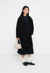 Zwarte wolmix jas met een hoge kraag, lange mouwen en rechte snit, gedragen over een marineblauw shirt en zwarte geplooide rok. Houdt een lichtbeige handtas vast.