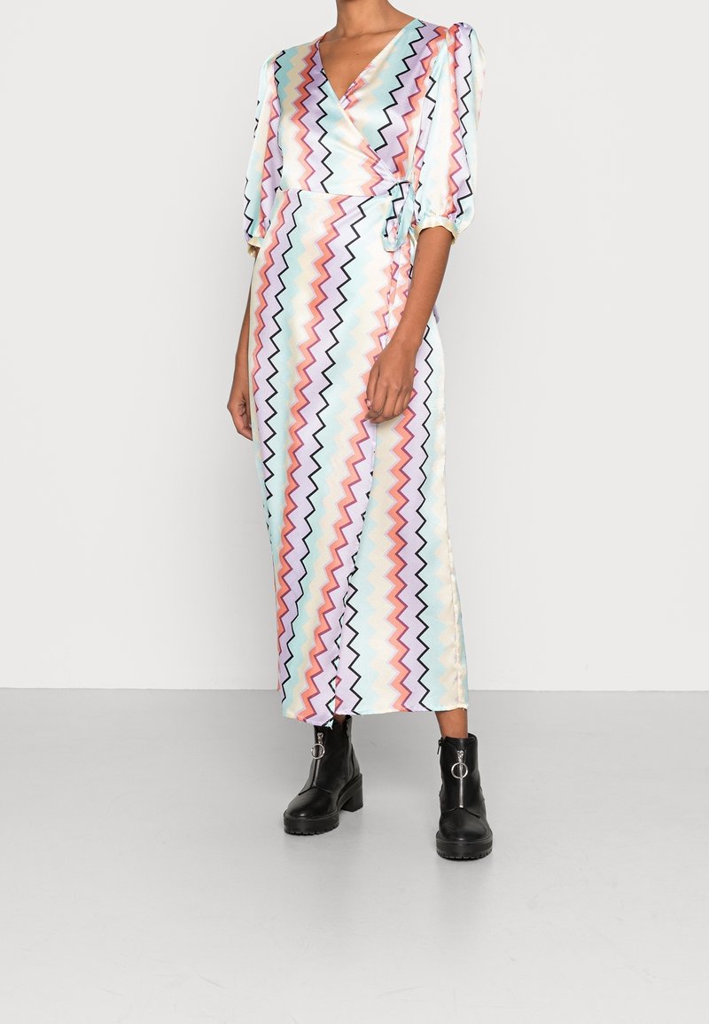 Lång, omslagsklänning med ett flerfärgat zigzag-mönster på en ljus bakgrund. Har tre fjärdedels ärmar och knytmidja, kombinerad med svarta stövlar.