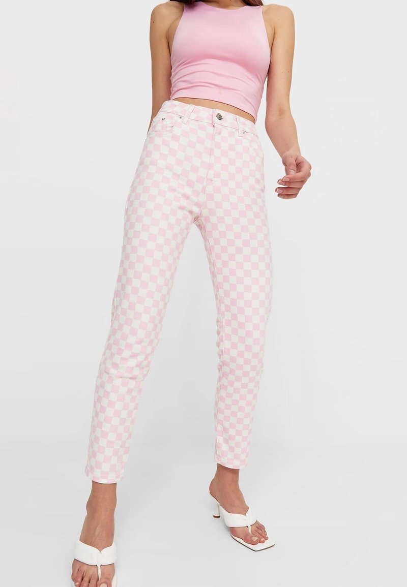 Femme portant un pantalon à carreaux rose et blanc, un top court sans manches rose et des sandales blanches à talons, sur un fond uni.