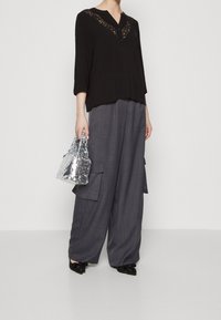 Frau in einem schwarzen Oberteil mit Spitzenbesatz, grauen weiten Cargohosen, schwarzen spitzen Schuhen und einer kleinen silbernen Pailletten-Handtasche.