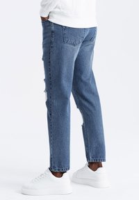 Vaqueros de denim azul claro con un corte relajado, que presentan detalles desgastados y dos bolsillos traseros, combinados con zapatillas blancas.