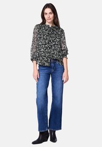 Schwarze transparente Bluse mit weißem Blumenmuster, Rüschenausschnitt und gepufften Ärmeln, kombiniert mit blauen Schlagjeans und schwarzen Stiefeletten.